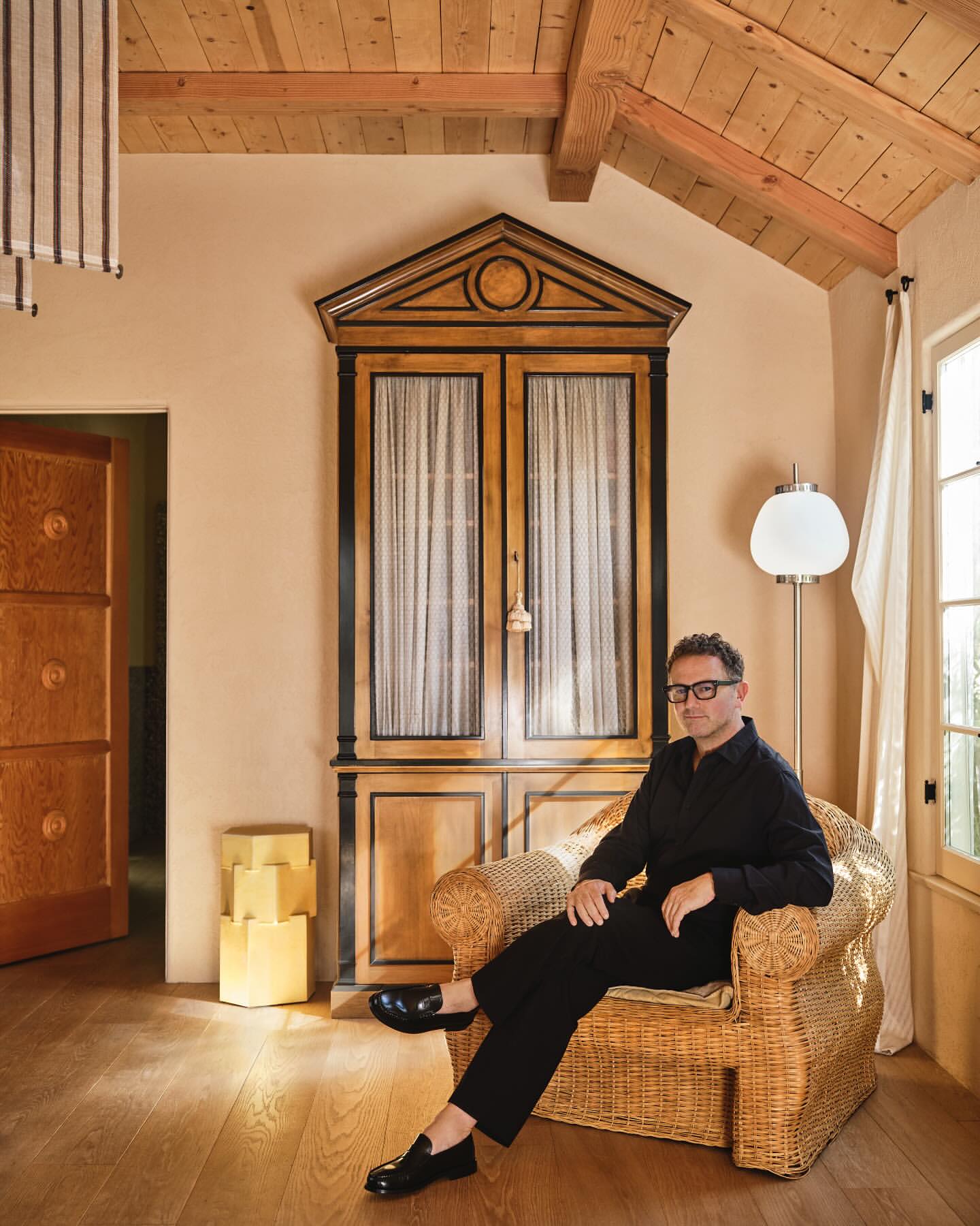 Adult male in black clothing sitting in a wicker chair in a home interior with neutral wall color, wooden floors and vintage decor.