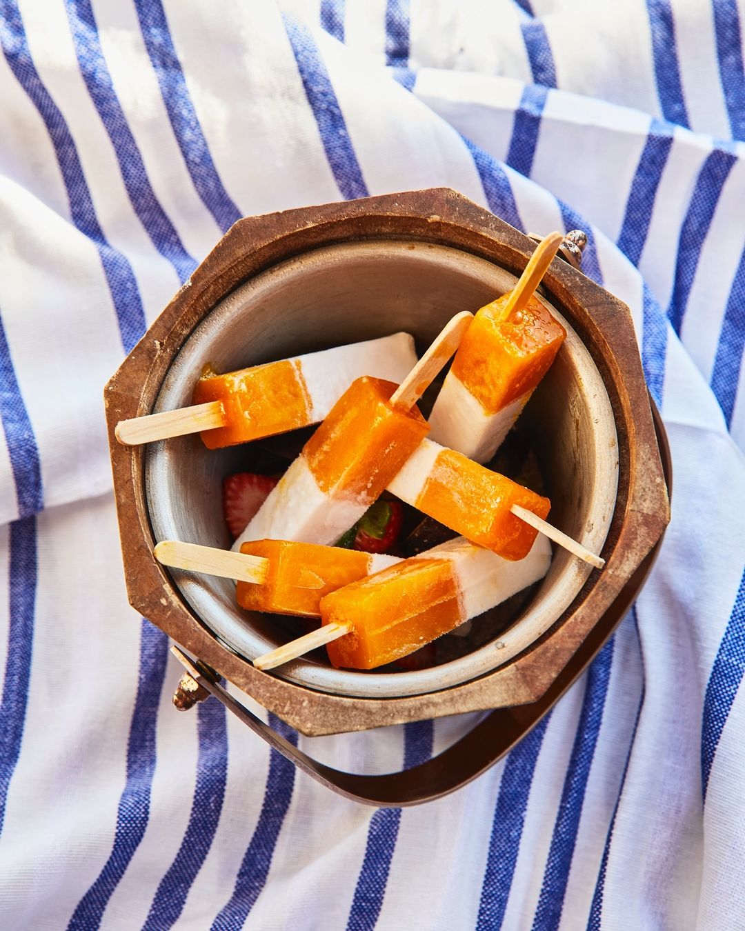 Overhead shot of a metal ice bucket filled with orange and white popsicles, along with strawberries, set on a striped towel.