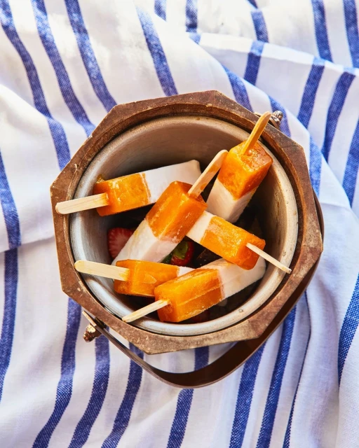 Overhead shot of a metal ice bucket filled with orange and white popsicles, along with strawberries, set on a striped towel.