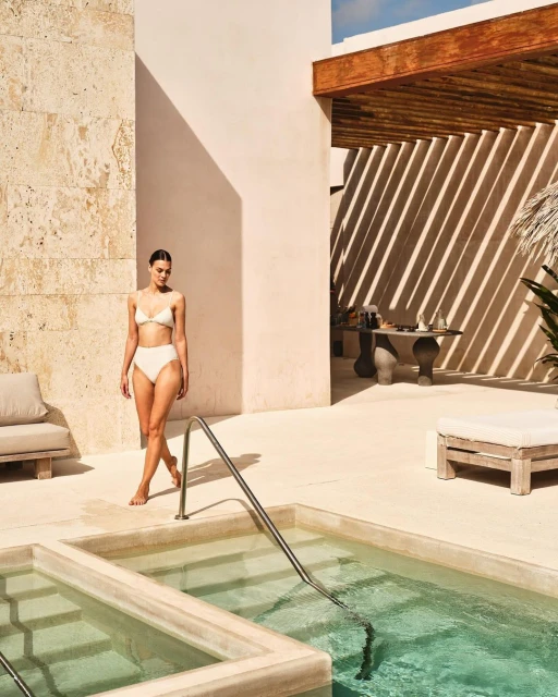 A woman in a swimsuit walking near a tiled pool surrounded by stone walls and lounge chairs in a resort setting with dappled light.