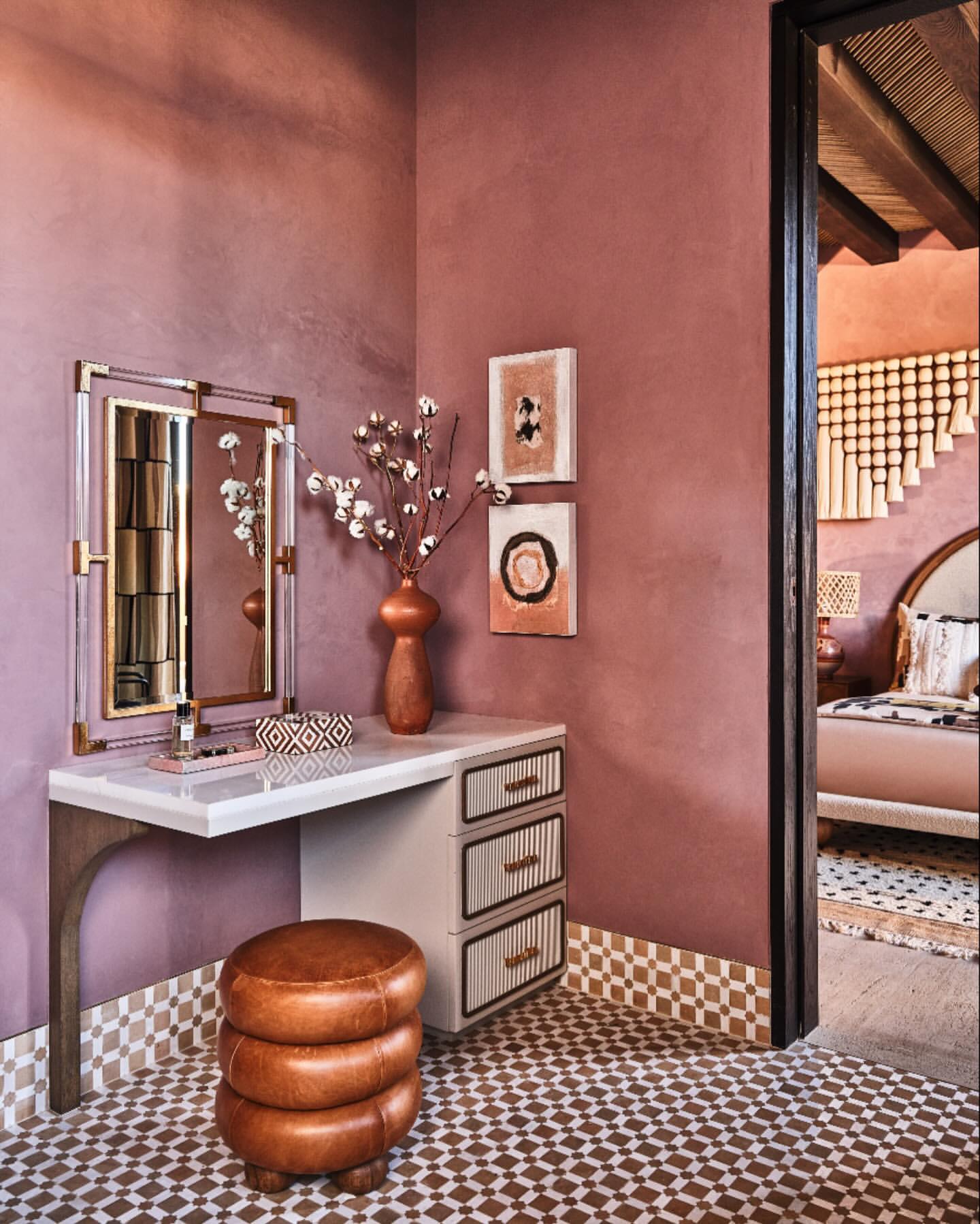 Interior design of a makeup vanity with mirror, flowers, stool, artwork, and geometric-patterned floor tiles in a room with mauve walls.