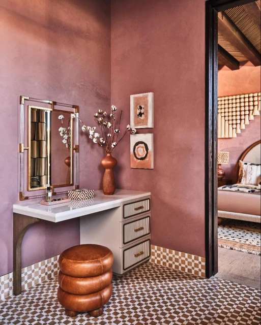 Interior design of a makeup vanity with mirror, flowers, stool, artwork, and geometric-patterned floor tiles in a room with mauve walls.