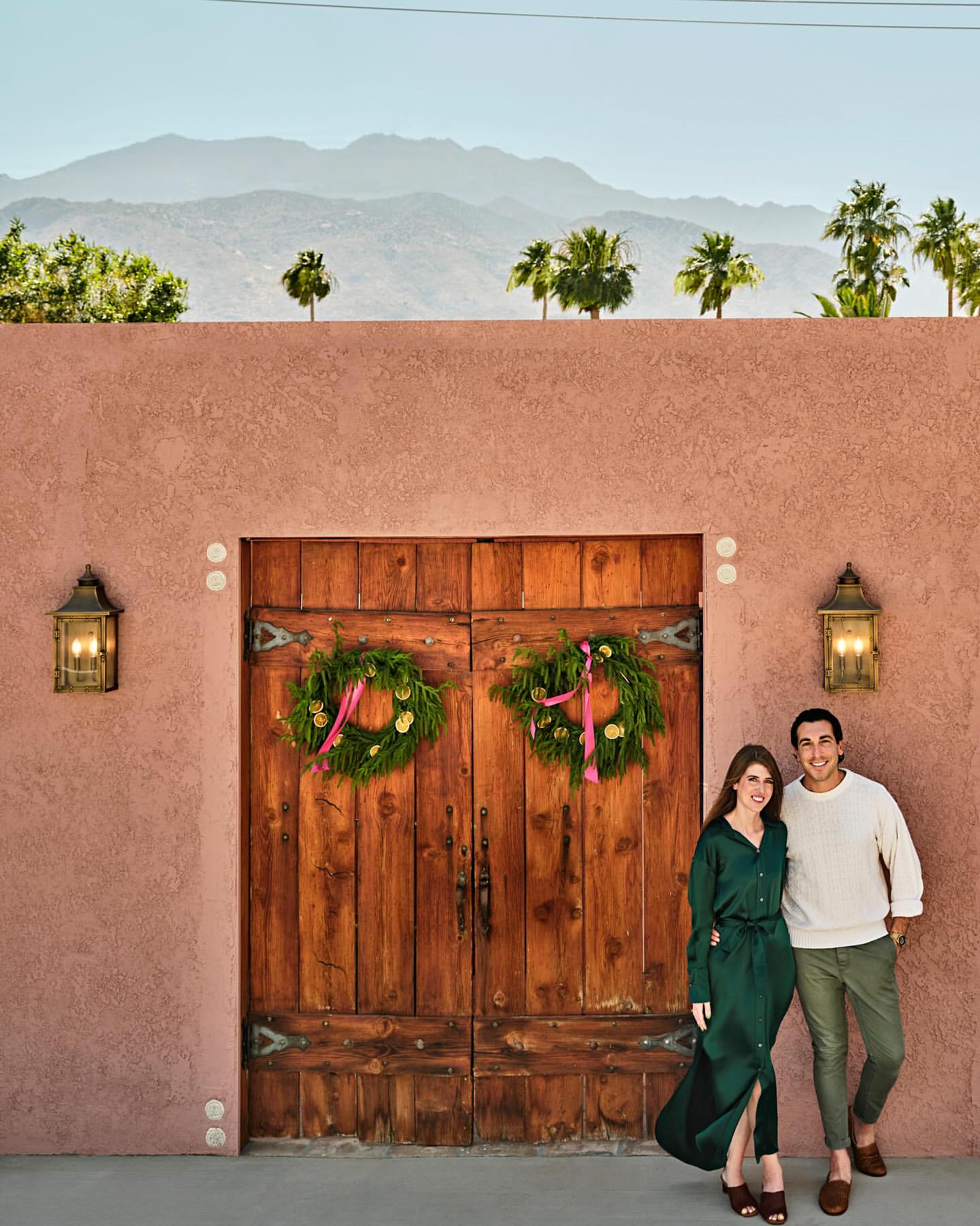 A happy couple stands in front of a rustic door adorned with Christmas wreaths, with mountains and palm trees in the background.