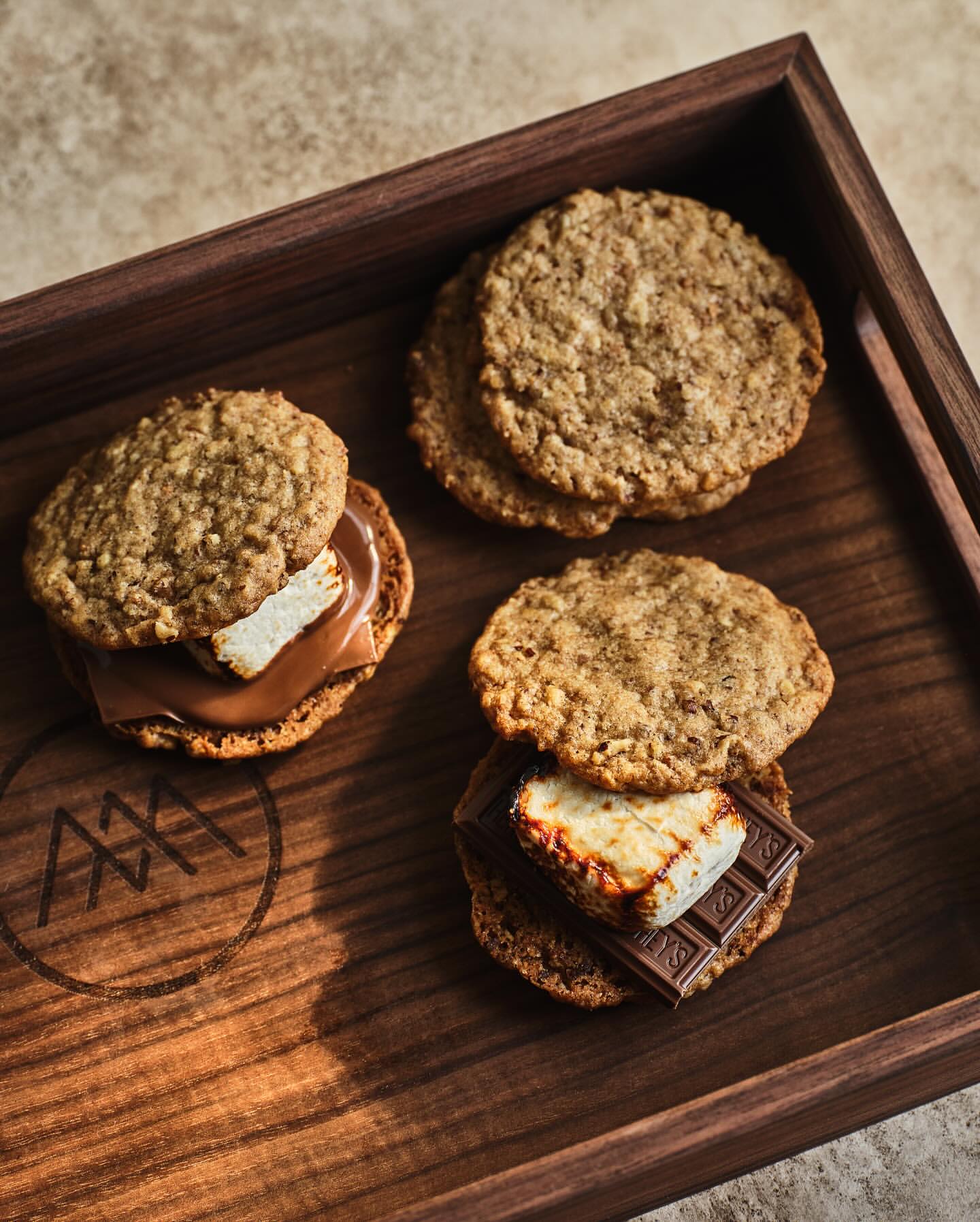 Gourmet smores with toasted marshmallows and chocolate between oat cookies, arranged on a wooden tray with logo detail.