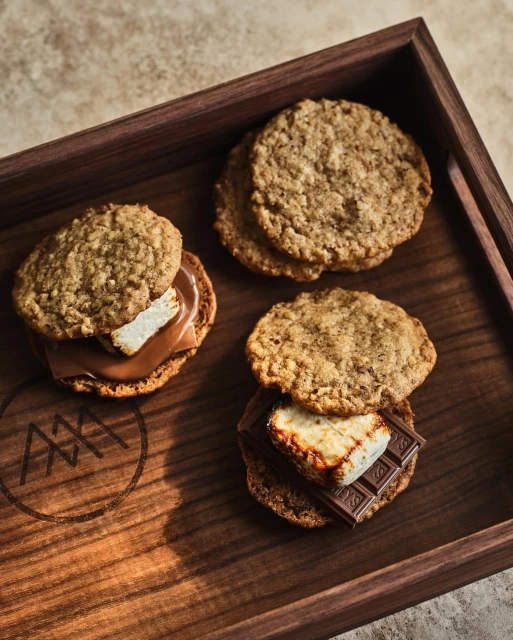 Gourmet smores with toasted marshmallows and chocolate between oat cookies, arranged on a wooden tray with logo detail.