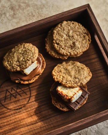 Gourmet smores with toasted marshmallows and chocolate between oat cookies, arranged on a wooden tray with logo detail.