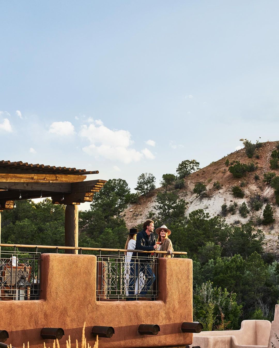 Three people standing on a stucco balcony with wooden beams, overlooking a hilly landscape under a pale sky, photographed by The Ingalls.