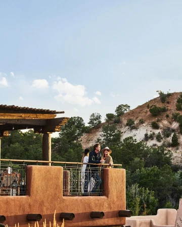 Three people standing on a stucco balcony with wooden beams, overlooking a hilly landscape under a pale sky, photographed by The Ingalls.