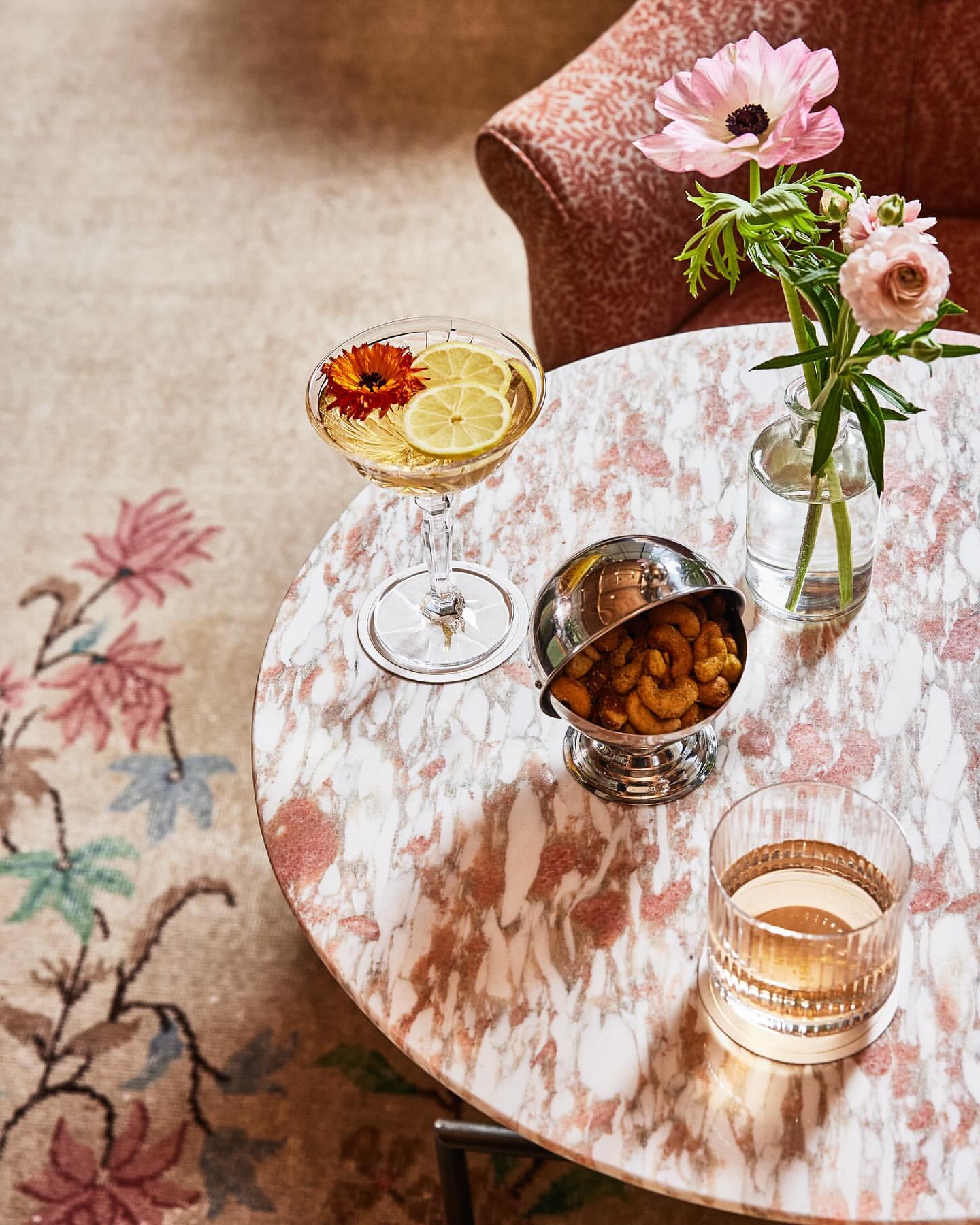 Overhead shot of a cocktail, snacks, and flowers on a pink marble table, creating an inviting and stylish atmosphere.