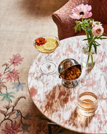 Overhead shot of a cocktail, snacks, and flowers on a pink marble table, creating an inviting and stylish atmosphere.
