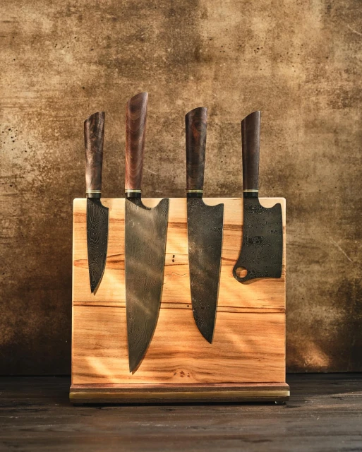 Four Damascus steel kitchen knives with wooden handles are displayed in a light-wood knife block against a mottled brown background.