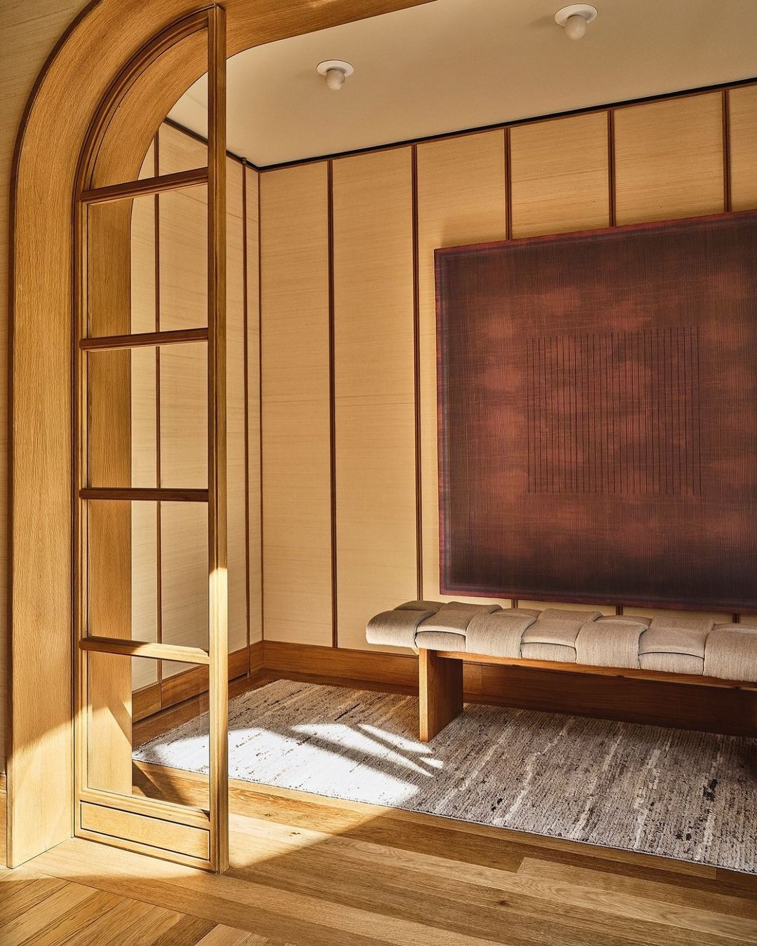 Indoor architectural shot of a wooden doorway, light beige paneled walls, a brown and beige woven bench, and an abstract art print.