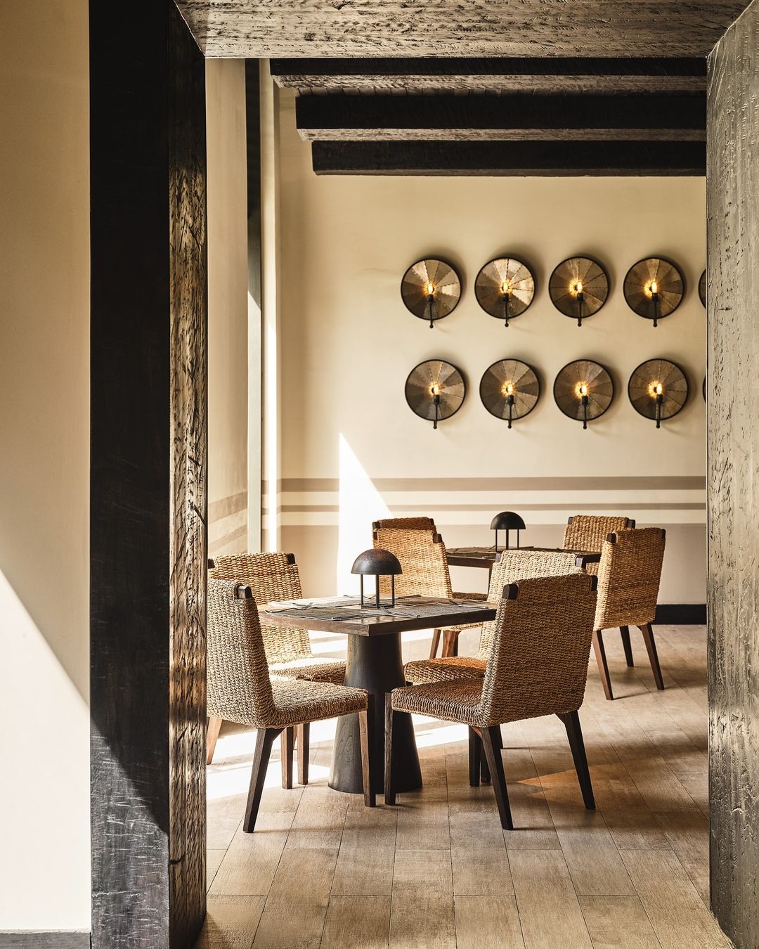 Indoor dining area featuring a wooden table and woven chairs, illuminated by sconce lighting on neutral walls and sunlight streaming through.