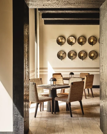 Indoor dining area featuring a wooden table and woven chairs, illuminated by sconce lighting on neutral walls and sunlight streaming through.