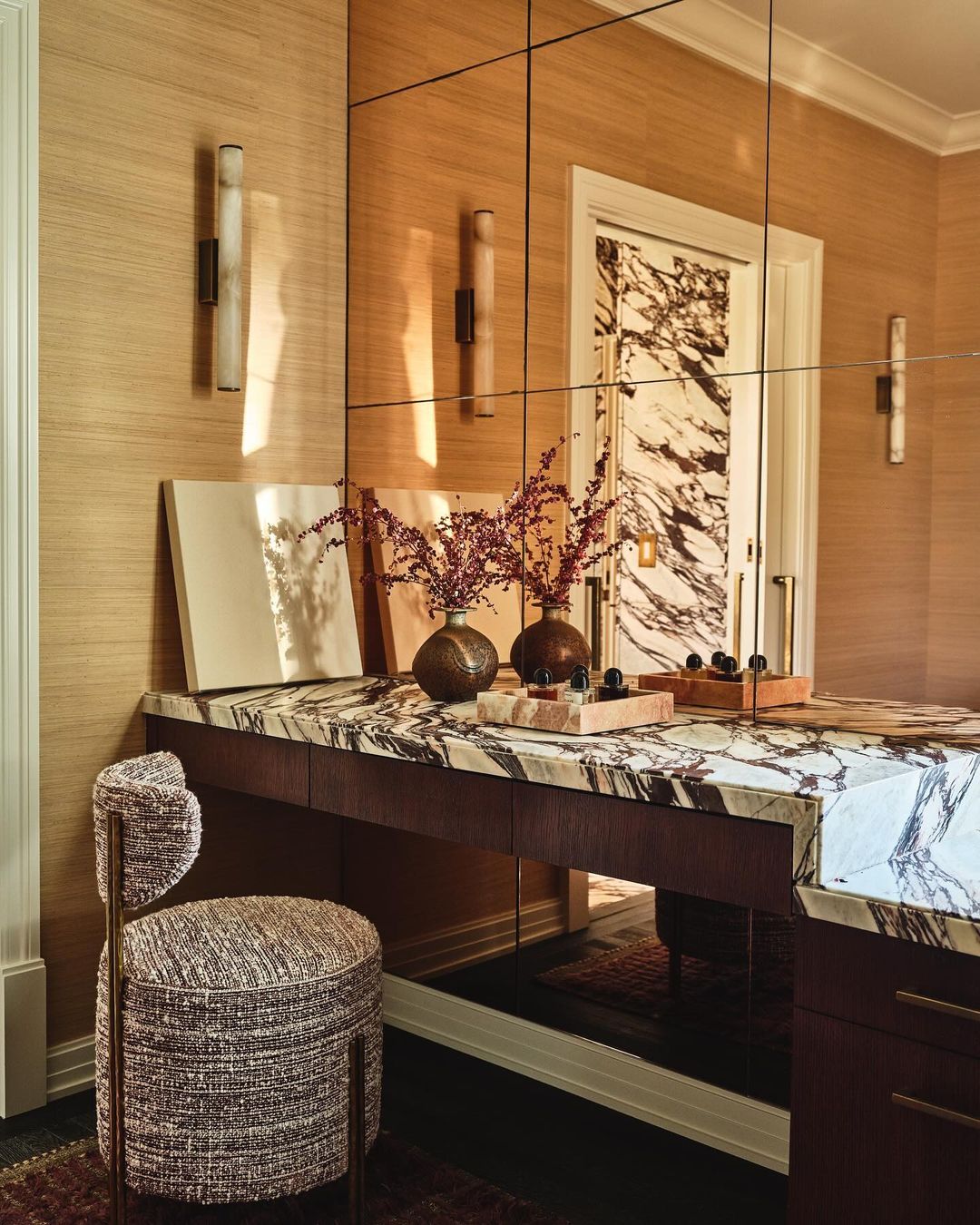 Interior design featuring a marble-topped vanity with floral arrangements, sconce lighting, decorative wall mirror, and a matching upholstered chair.