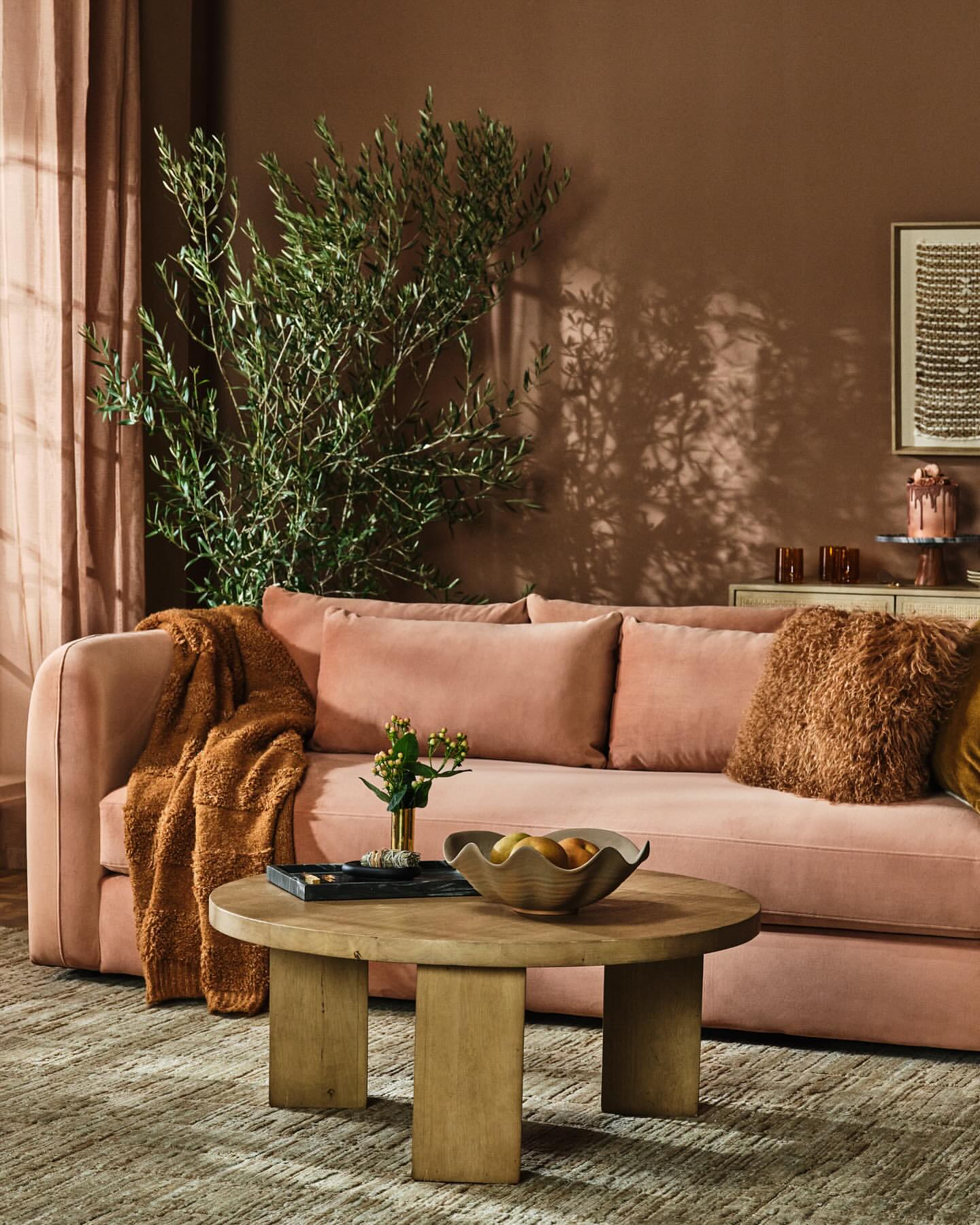 Living room interior showcasing a pink sofa with pillows, a wooden coffee table with fruit, and olive tree branches, creating a cozy ambiance.