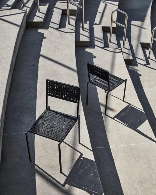 Two black woven chairs sit on a concrete surface with long, linear shadows in a minimalist, high-angle architectural composition.