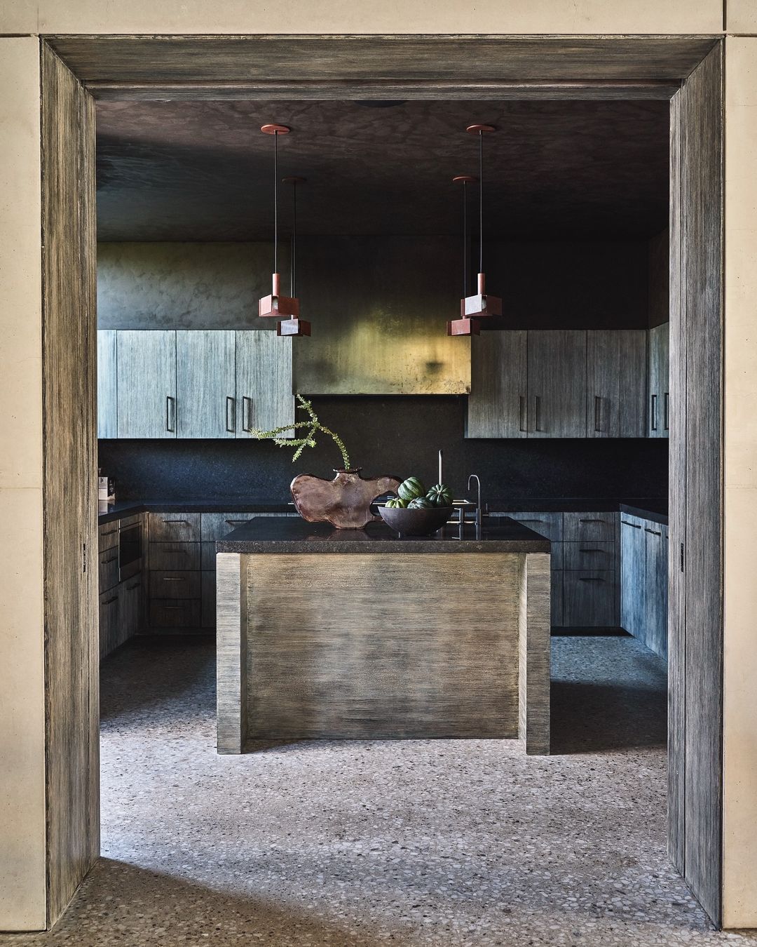 Modern kitchen interior with gray wood cabinets, black countertops, island with bowl of green artichokes, pendant lighting and textured floor.