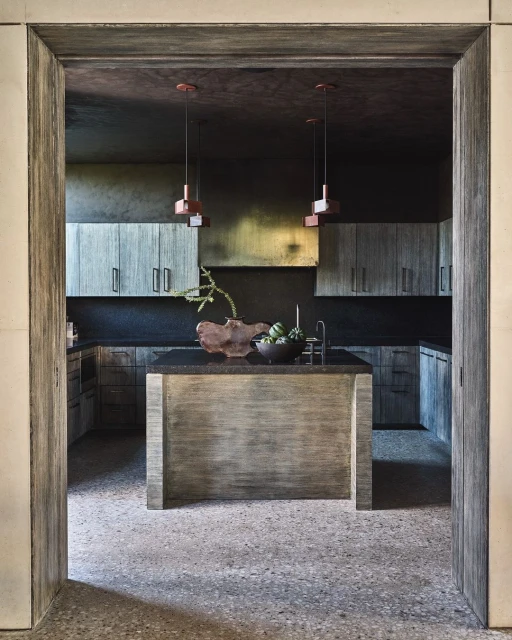 Modern kitchen interior with gray wood cabinets, black countertops, island with bowl of green artichokes, pendant lighting and textured floor.