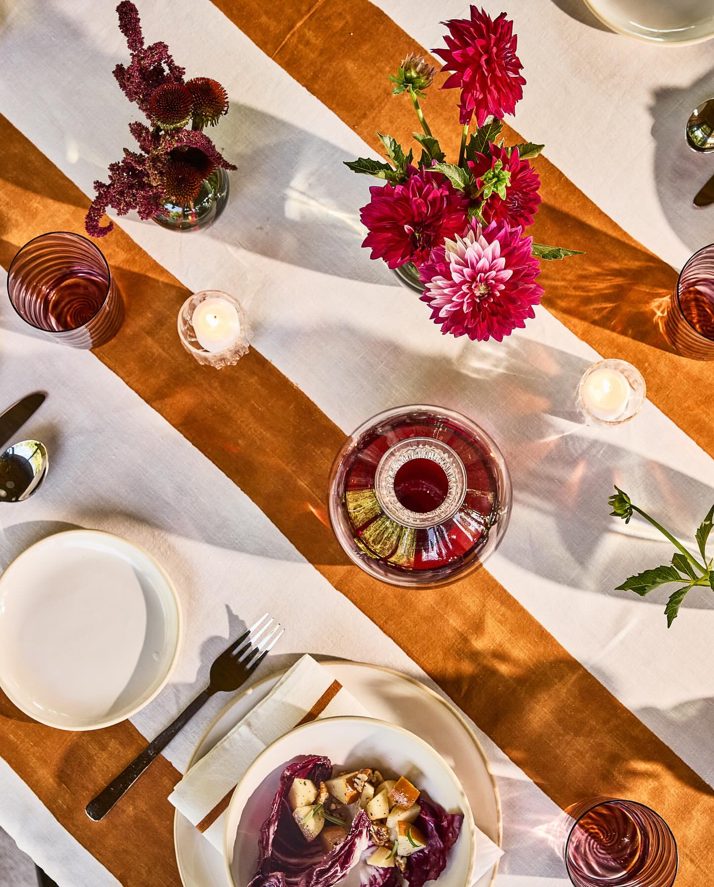 Overhead shot of a dining table with floral centerpieces, candles, salad plates, silverware, and striped linen tablecloth with warm lighting.