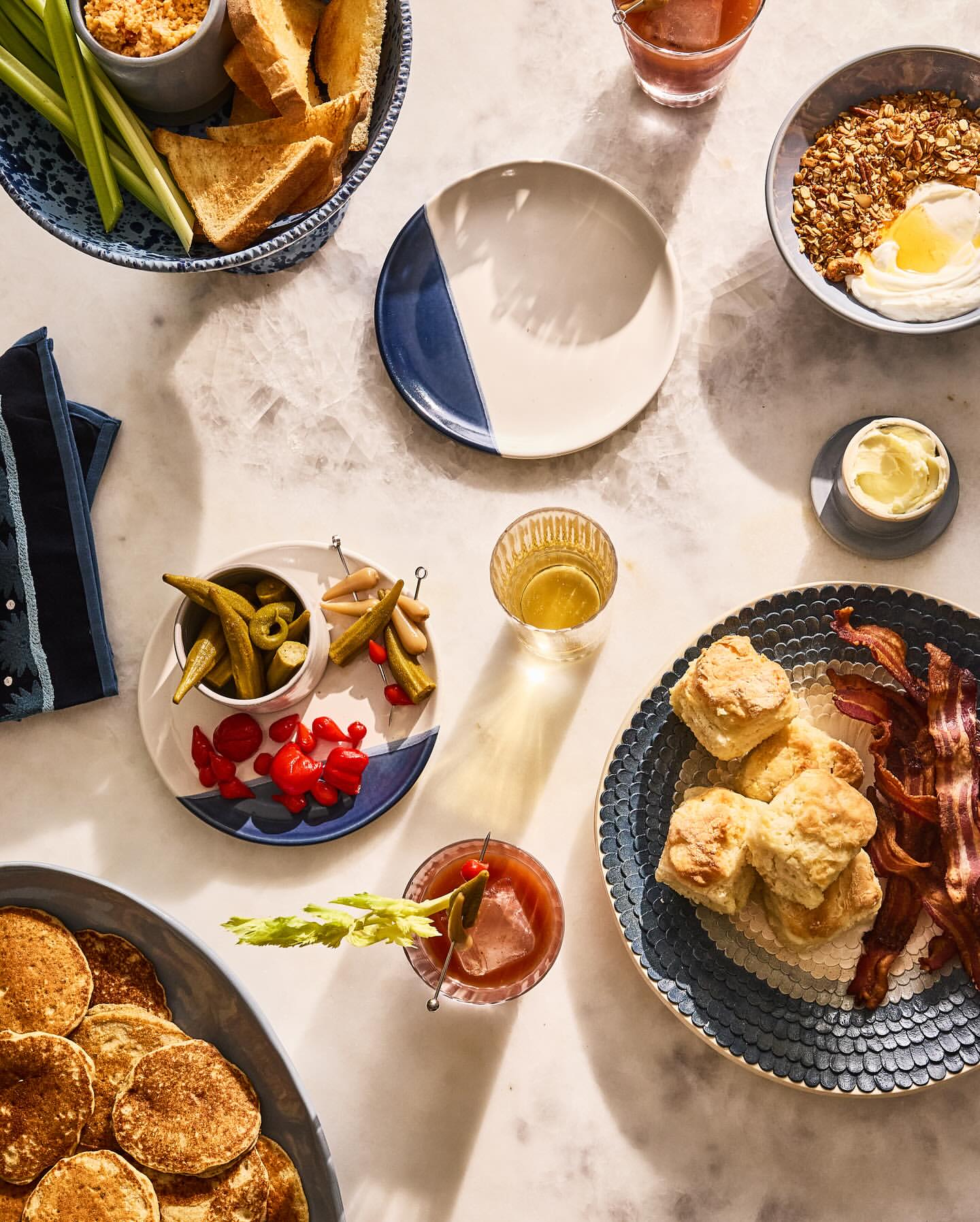 Overhead shot of breakfast food including pancakes, biscuits, bacon, grits, celery sticks, toast, and cocktails on a marble table.