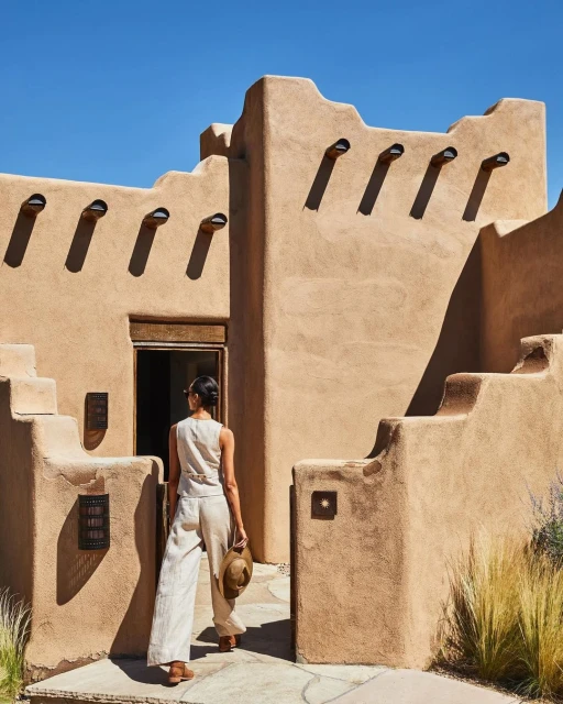 A woman in casual linen clothing walks toward an adobe-style building under a clear blue sky, holding a straw hat in hand.