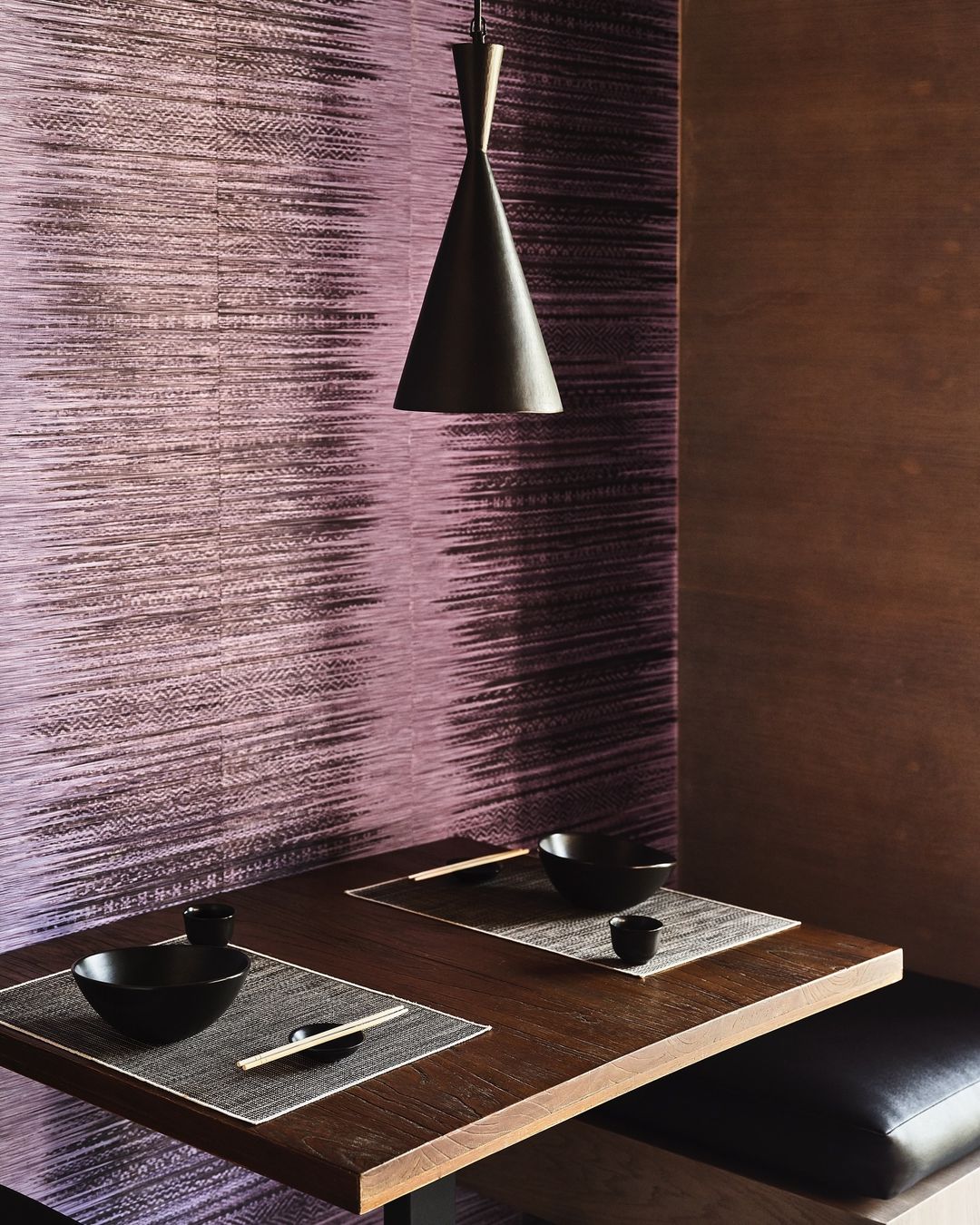 Dining nook with dark wood table, black bowls and placemats, illuminated by a single pendant light against a textured wall.