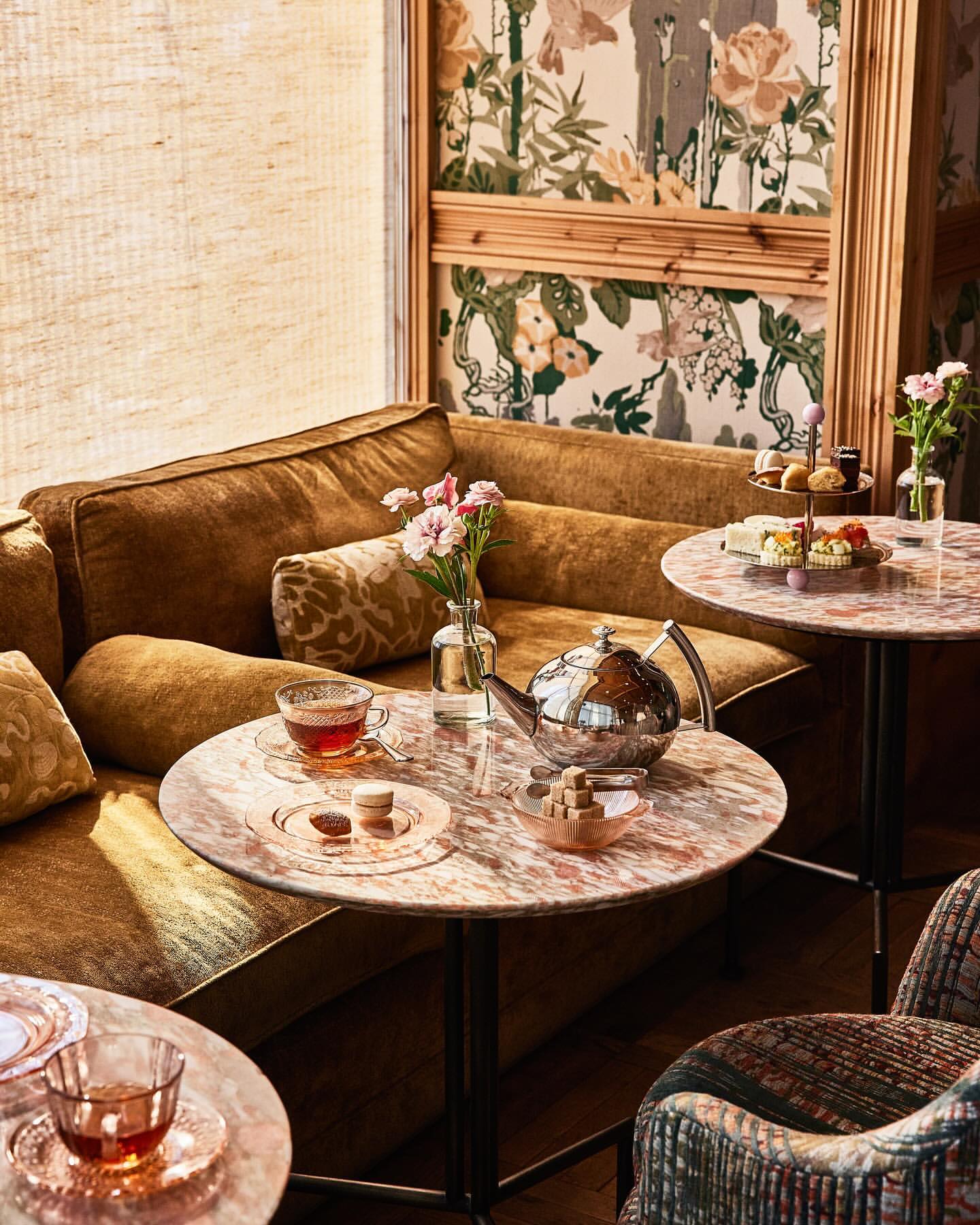 Tea service with a silver teapot, tea cups, and desserts set on marble-topped tables in an elegant lounge area with floral wallpaper.