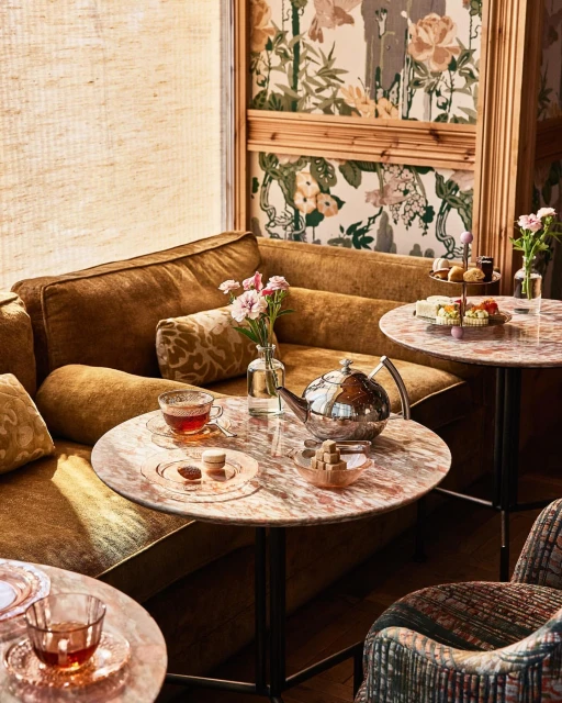 Tea service with a silver teapot, tea cups, and desserts set on marble-topped tables in an elegant lounge area with floral wallpaper.