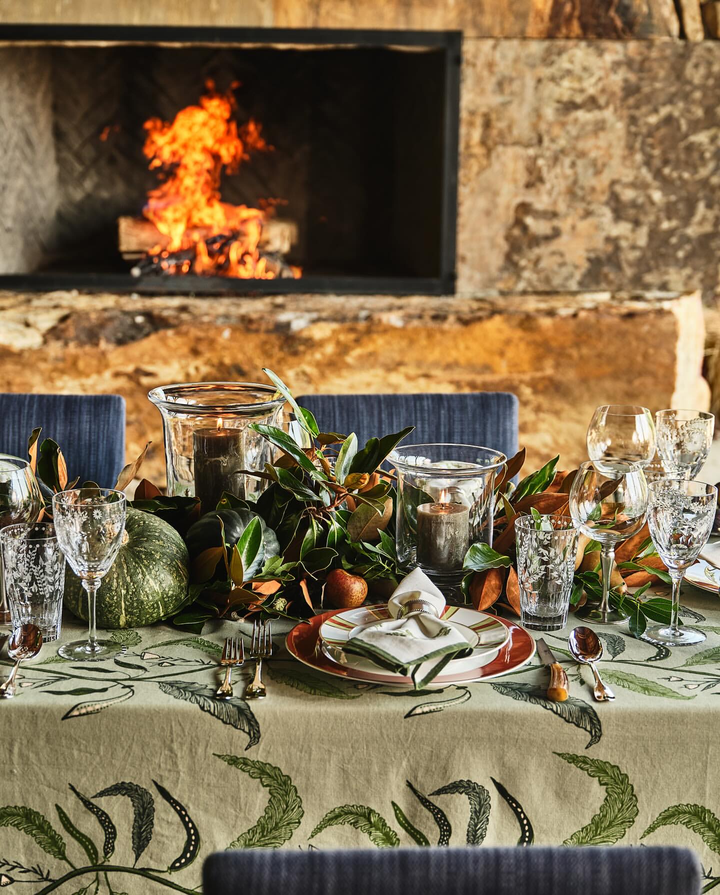 Festive table setting with candles, greenery, plates, glassware, and pumpkins in front of an indoor stone fireplace. Cozy atmosphere.