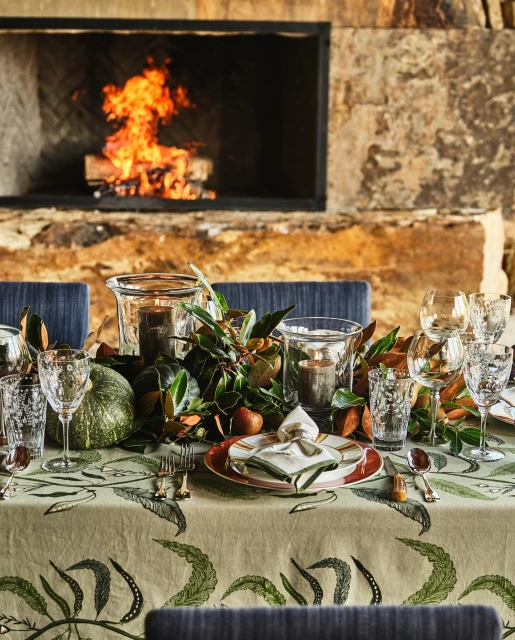 Festive table setting with candles, greenery, plates, glassware, and pumpkins in front of an indoor stone fireplace. Cozy atmosphere.