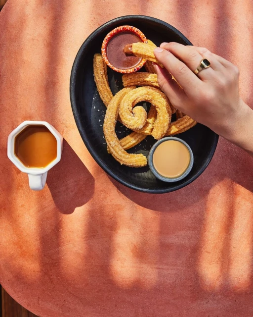 Overhead shot of churros on a black plate with dipping sauces, a cup of coffee, and a hand holding a churro.