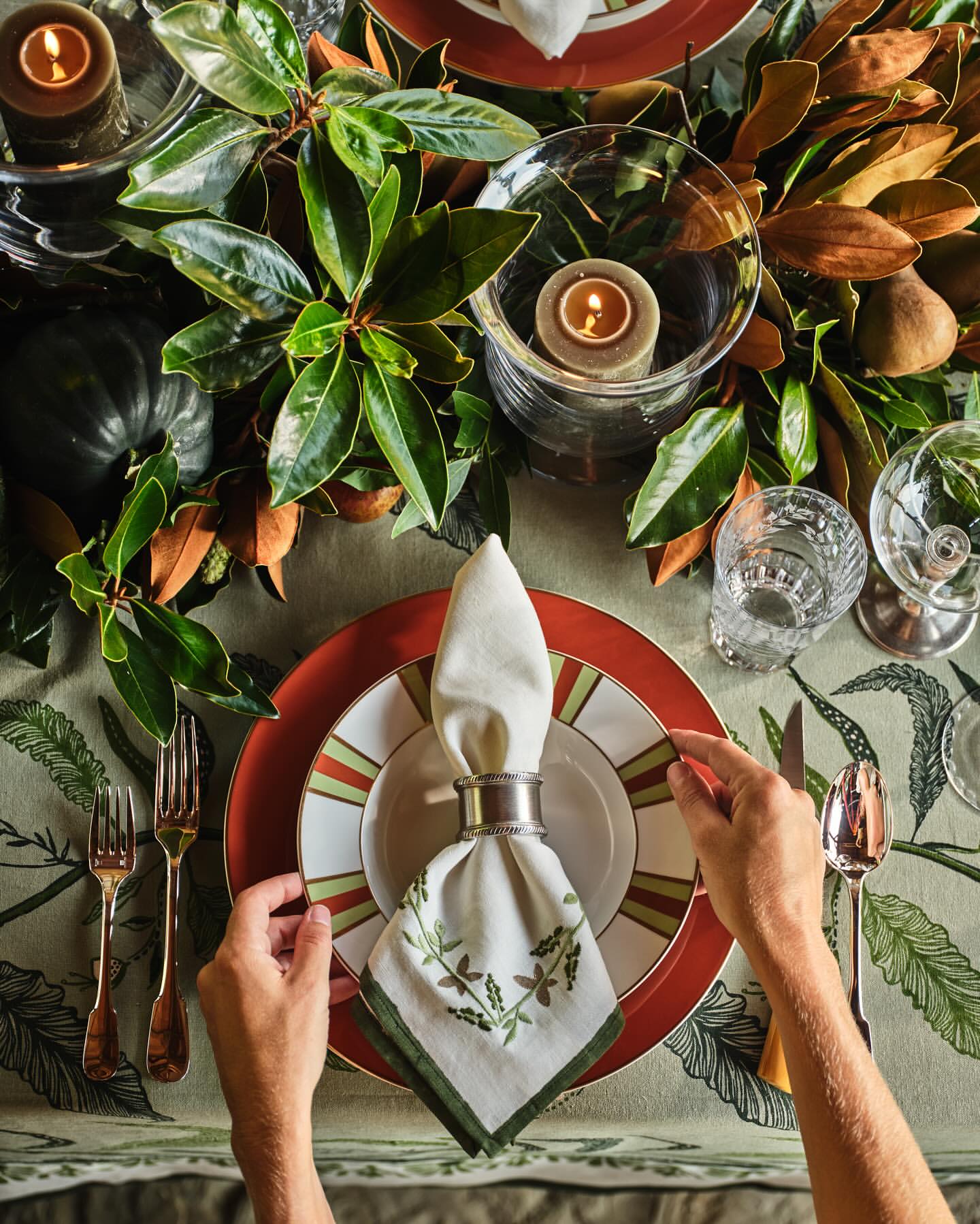 Overhead shot of a decorated table setting featuring dinnerware, candles, leafy foliage, water glasses, silverware, and a table cloth.