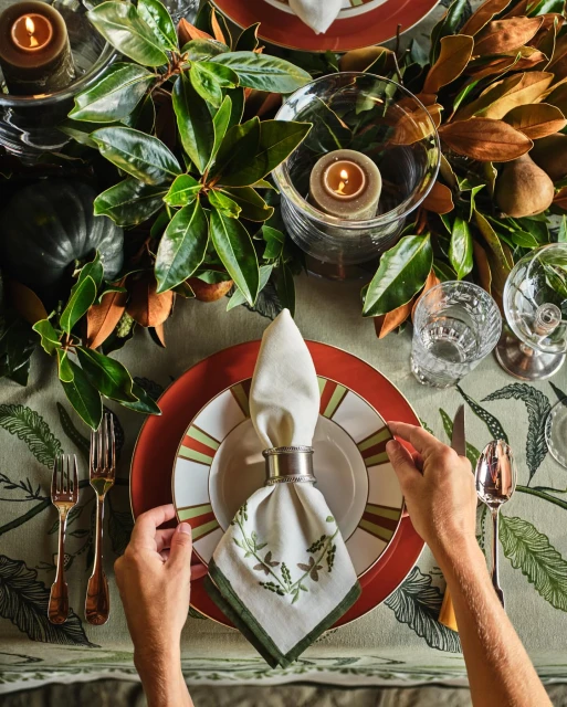 Overhead shot of a decorated table setting featuring dinnerware, candles, leafy foliage, water glasses, silverware, and a table cloth.