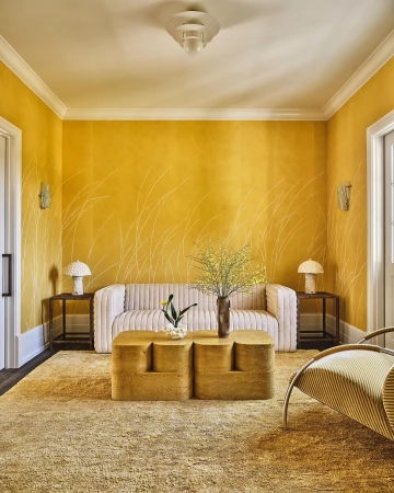 Interior view of a living room featuring a cream-colored sofa, geometric coffee table, area rug, and yellow walls with subtle decorations.