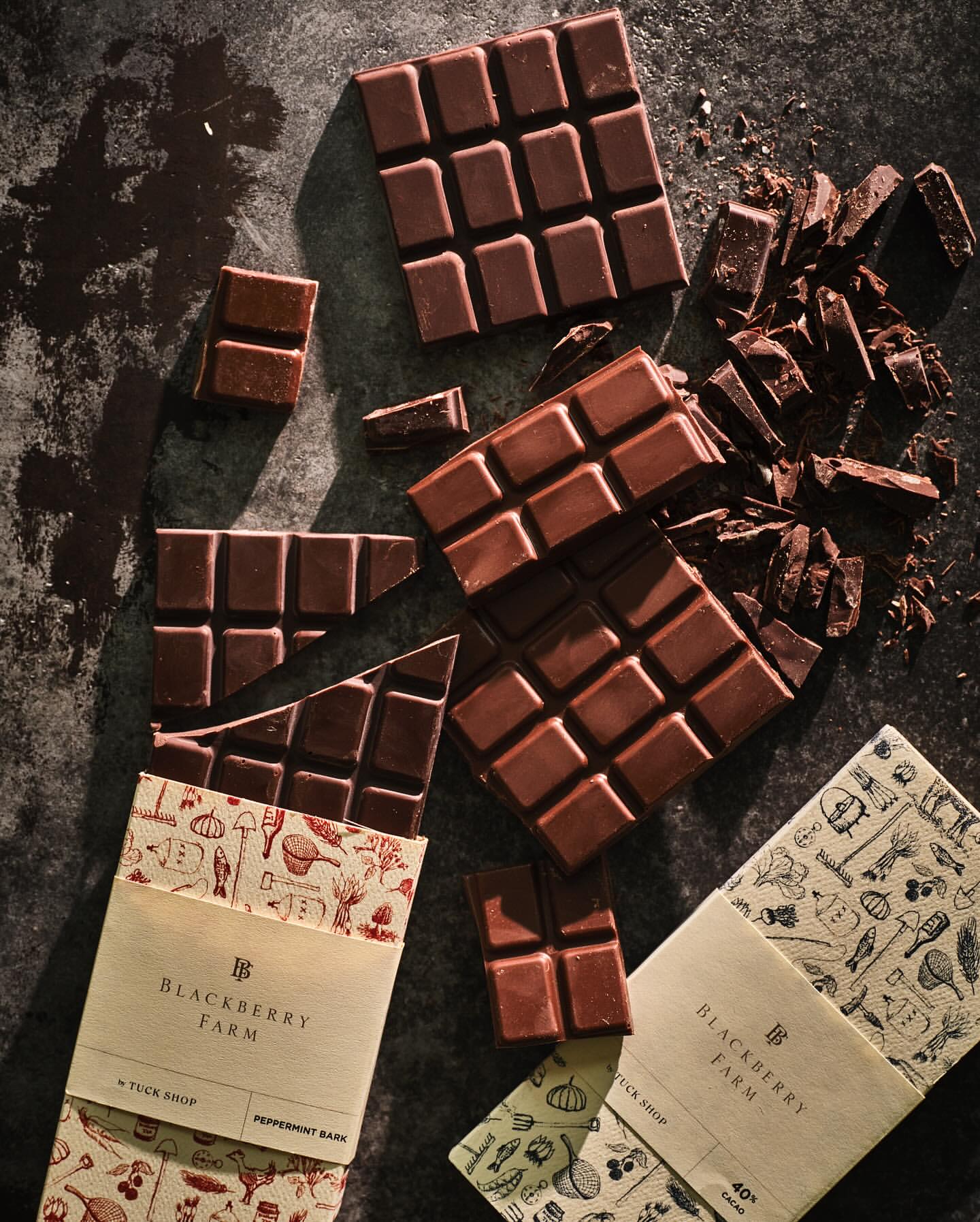 Overhead shot of broken chocolate bars with paper wrappers scattered on a dark surface, highlighting food photography and dark brown hues.