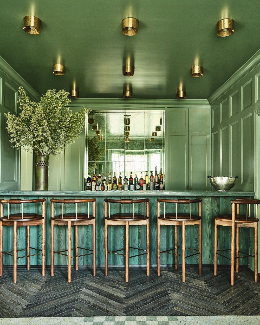 Interior view of a home bar with pale green walls, wooden stools, mirrored bar, and display of liquor bottles, enhancing a sophisticated space.