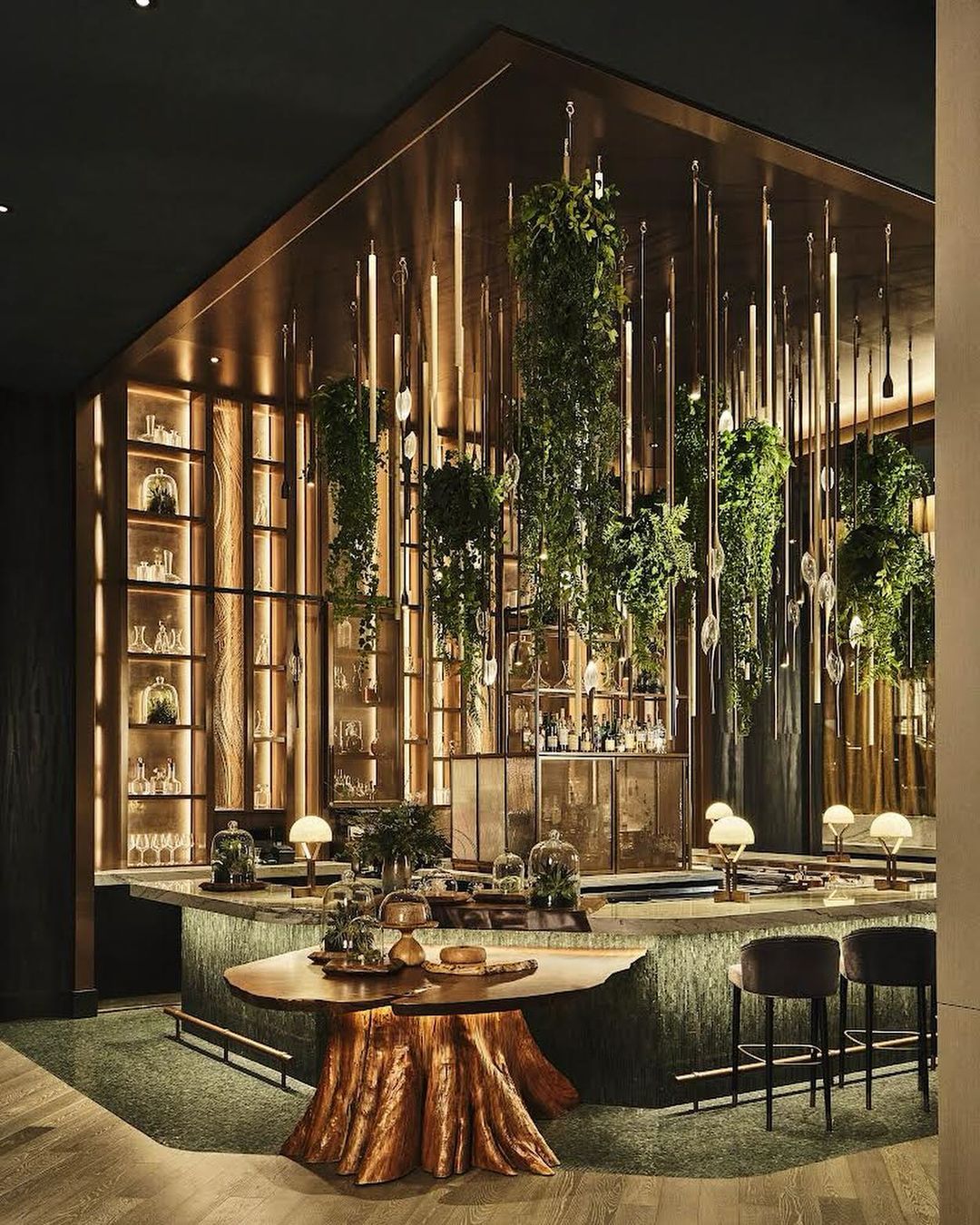 Interior of an upscale bar area featuring a unique bar table and an elaborate display of hanging plants and artistic lighting fixtures.