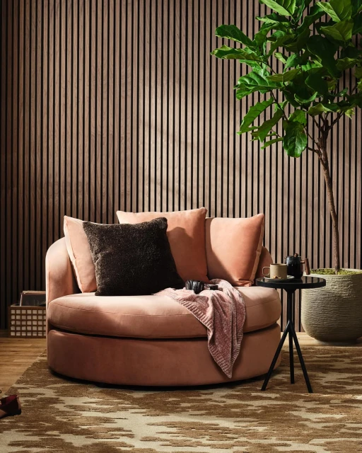 Interior with a round peach-colored sofa, throw pillows, blanket, side table with tea set, fig tree, and brown wood paneling wall.