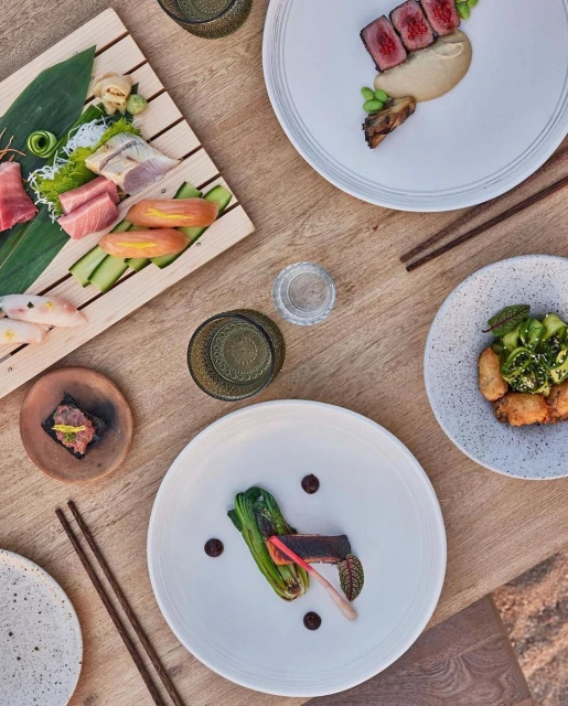Overhead shot of a wooden table featuring an assortment of beautifully plated dishes, including sushi and meat, accompanied by chopsticks and drinks.
