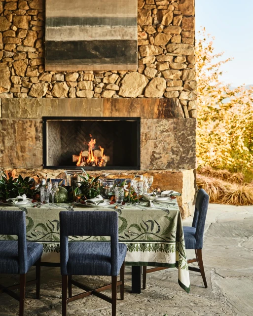 Outdoor dining table is set next to a stone fireplace with burning logs, with blue upholstered chairs and a decorative green tablecloth.