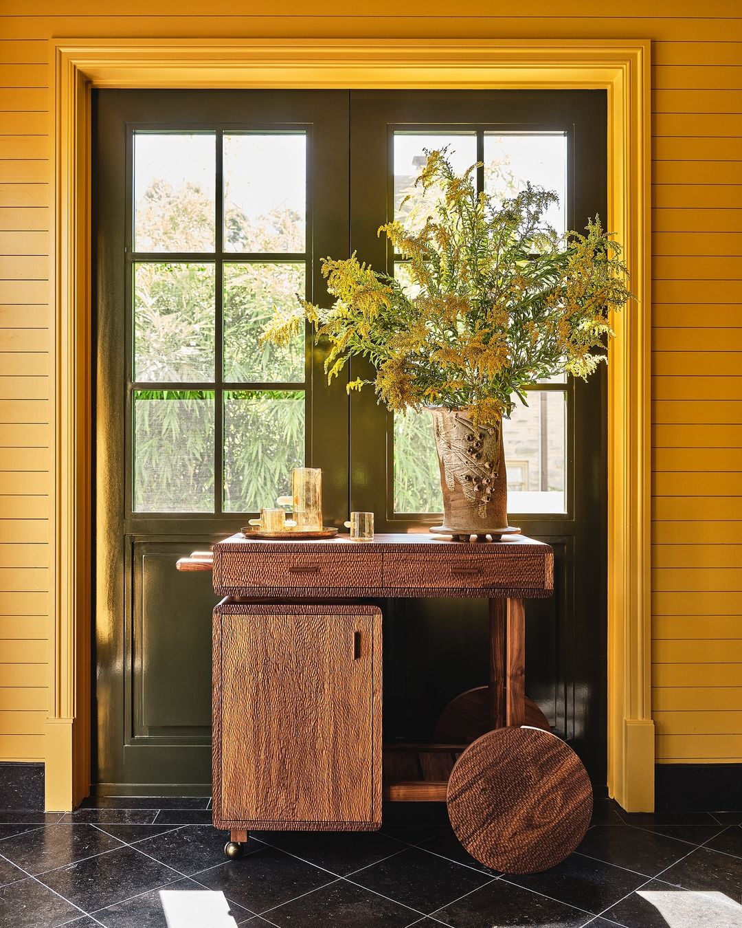Indoor shot of a decorative bar cart with a floral arrangement, set against a backdrop of a window and yellow horizontal panelling.