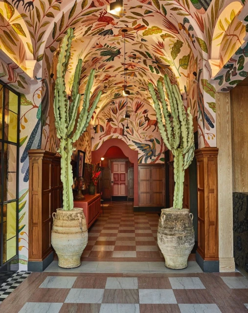 Interior view of a corridor with a checkered floor, arched ceiling covered in a leaf and animal pattern, and potted cacti.