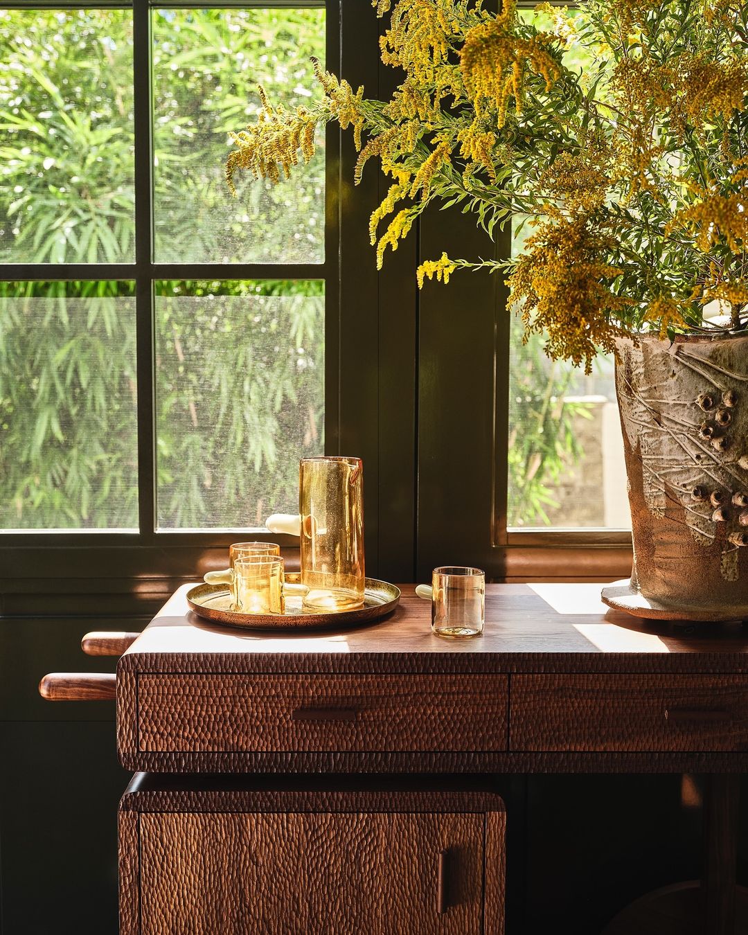 Amber glassware and a brass tray on a wooden textured console table with a large vase of yellow flowers.