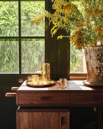 Amber glassware and a brass tray on a wooden textured console table with a large vase of yellow flowers.