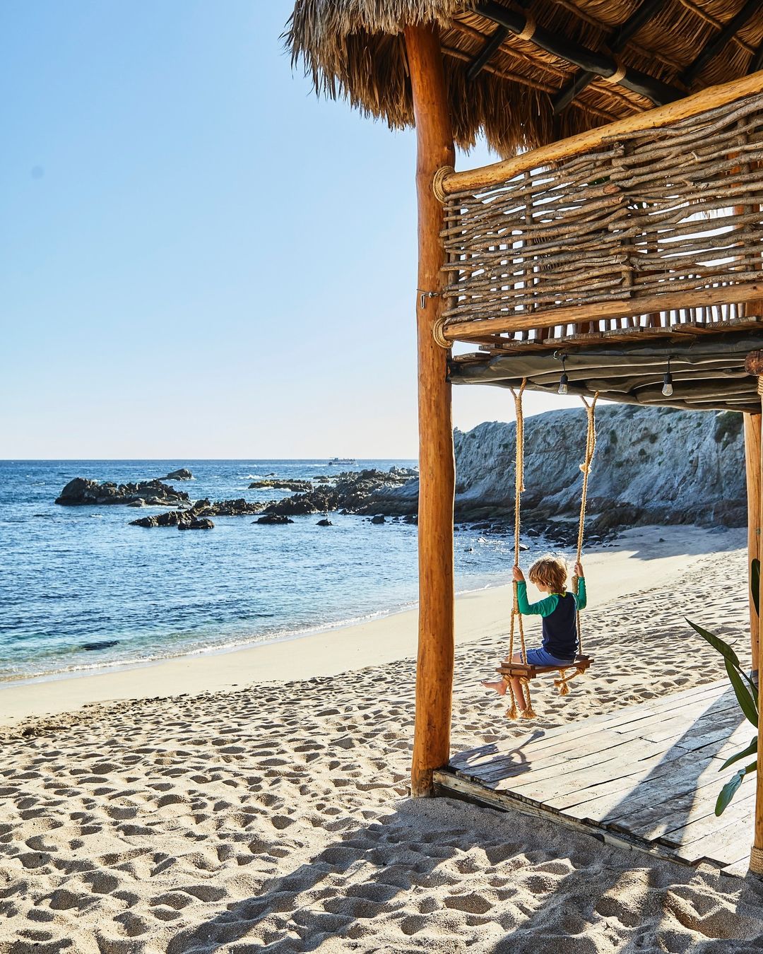 Child on a wooden swing at a beachside hut overlooking the ocean with white rocks, sand, and clear blue sky in a travel image.