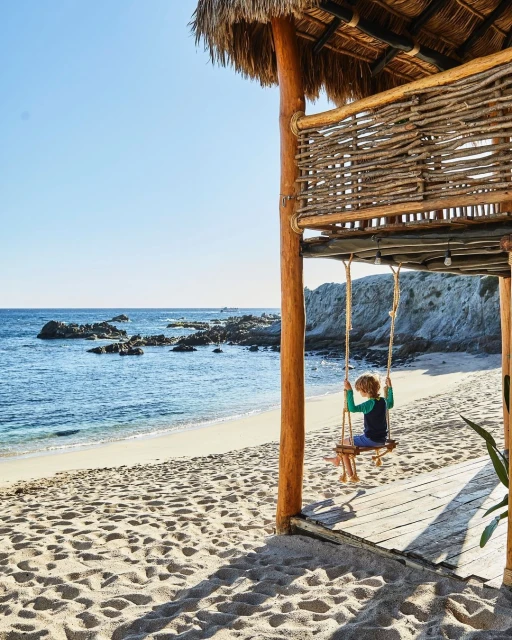 Child on a wooden swing at a beachside hut overlooking the ocean with white rocks, sand, and clear blue sky in a travel image.