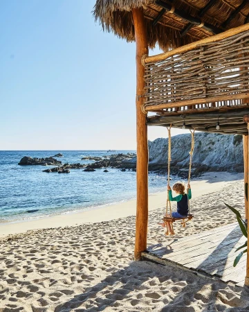 Child on a wooden swing at a beachside hut overlooking the ocean with white rocks, sand, and clear blue sky in a travel image.
