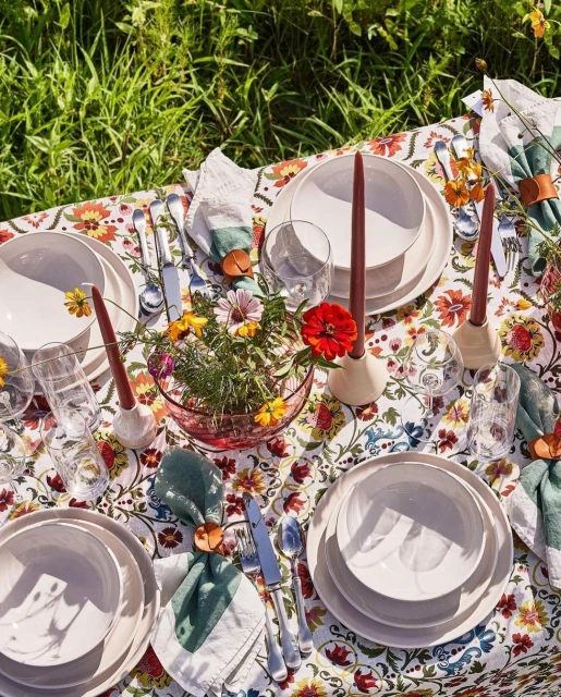 Overhead shot of an outdoor dining table set with floral tablecloth, white plates, flower-filled centerpiece, candles, and napkins.
