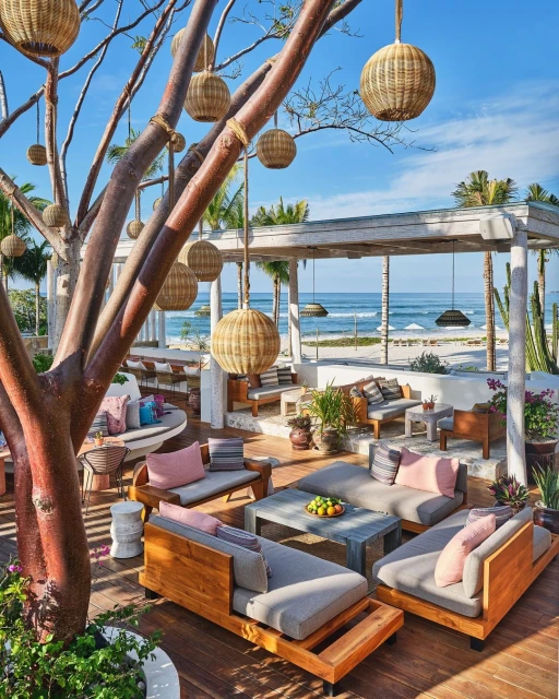 Outdoor lounge area at a tropical resort with cushioned seating, wooden furniture, and woven pendant lights under a clear blue sky near the ocean.